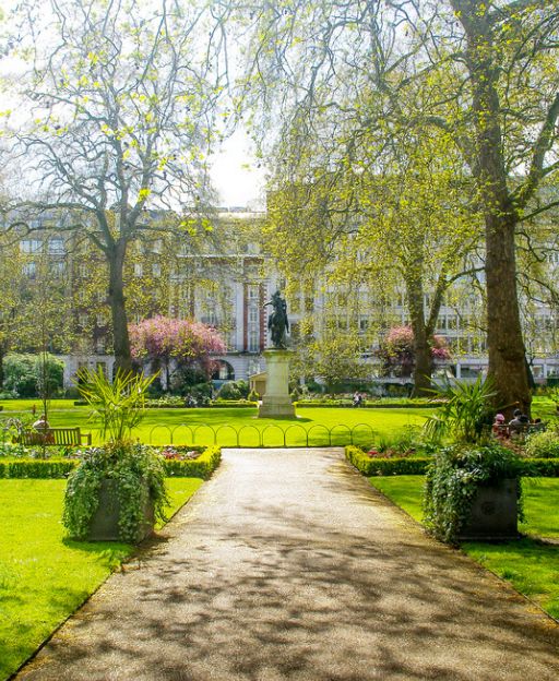 St James's Square Garden in central London, a formal garden with a gravel path, green lawns, a central statue, and a grand white building beyond spring trees
