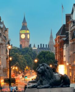 Trafalgar Square at dusk with Big Ben visible and a lion statue in the foreground, London