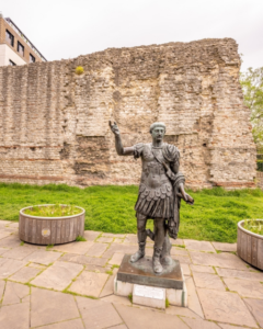 Bronze statue of Roman Emperor Trajan standing before a surviving section of ancient Londinium wall in the City of London