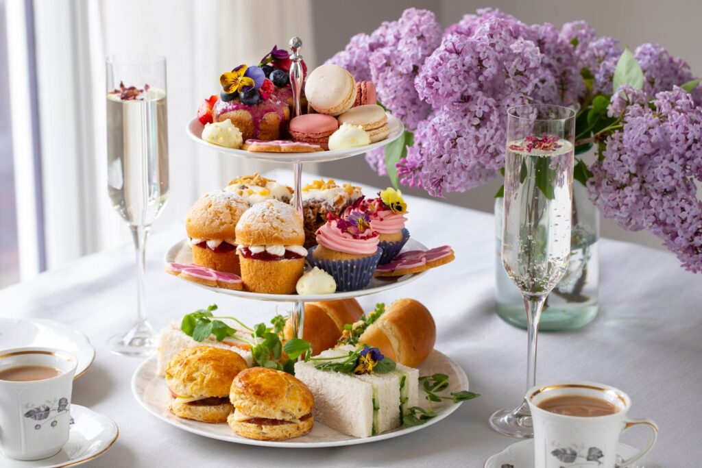 A traditional afternoon tea on a three-tier stand: finger sandwiches and scones at the bottom, cakes and macarons above, with cups of tea and glasses of sparkling wine