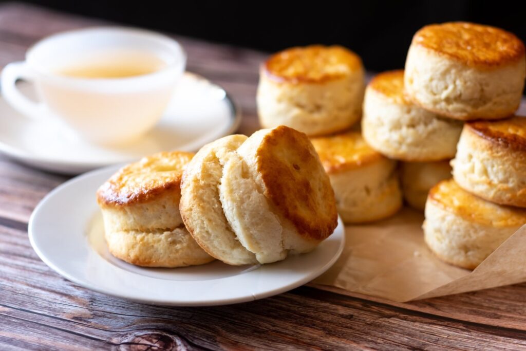 A pile of freshly baked golden plain scones on a plate beside a cup of tea