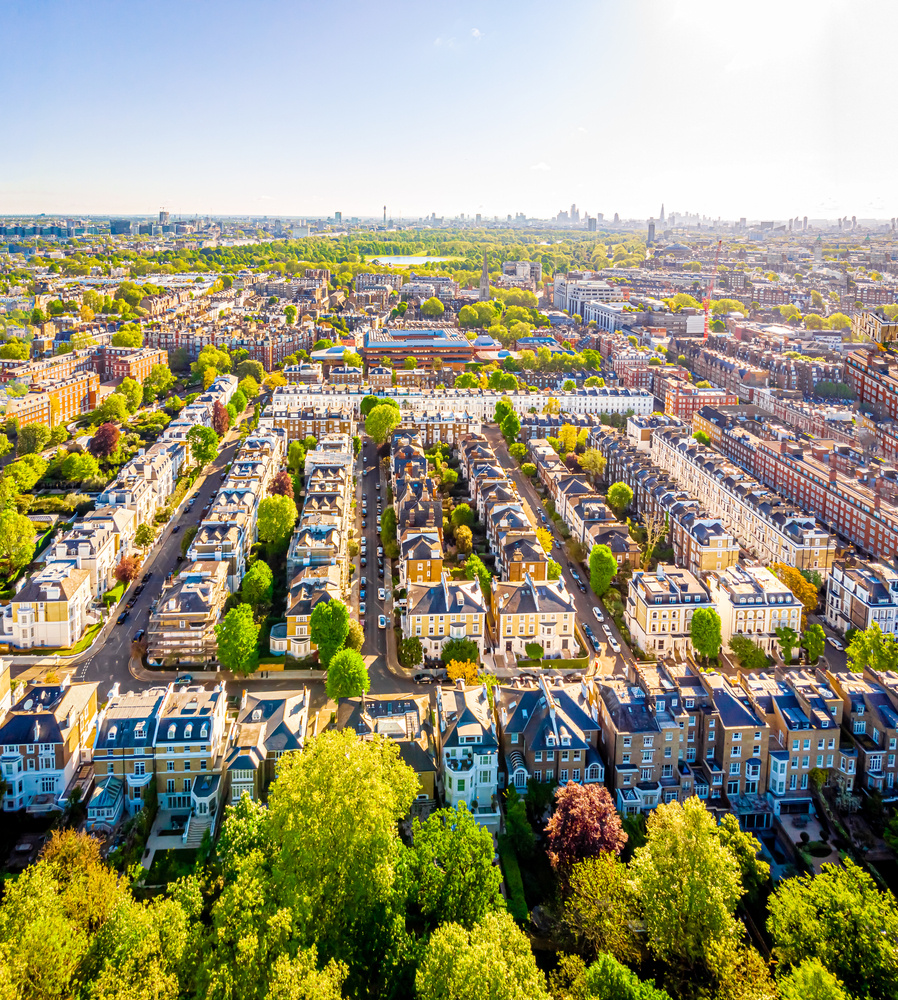 Aerial view of Kensington's cream terraces and green squares on a sunny morning, with the London skyline in the distance