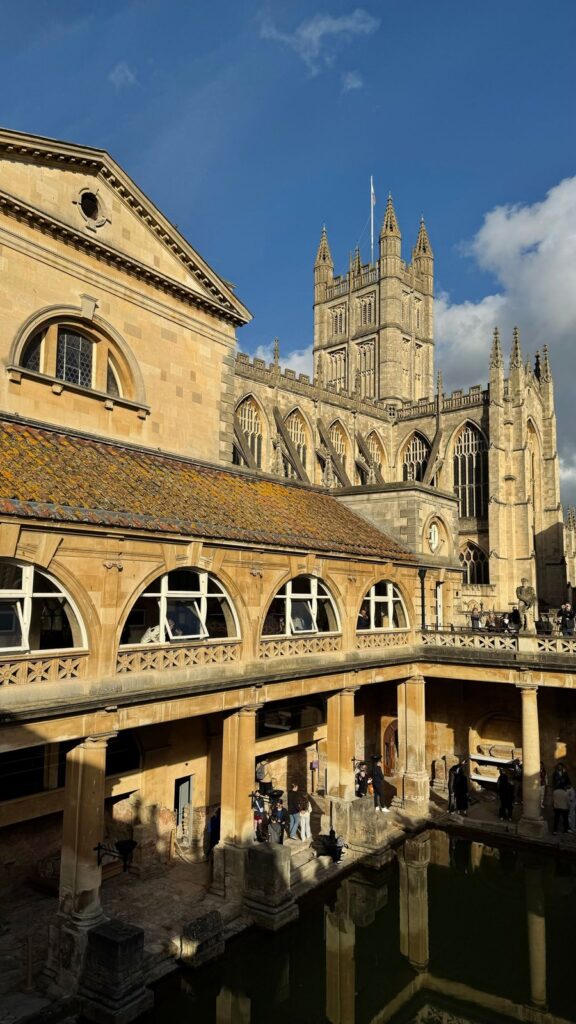 The Great Bath at the Roman Baths in Bath with Bath Abbey tower behind under a blue sky