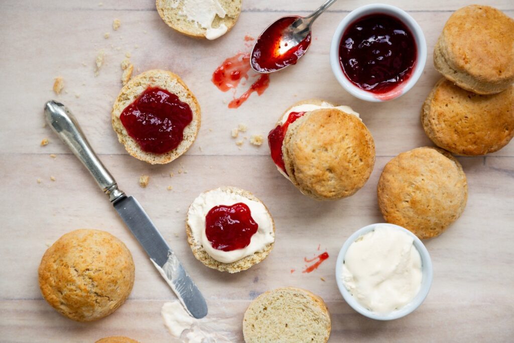 Halved English scones topped with clotted cream and strawberry jam, with small bowls of jam and cream on a wooden table